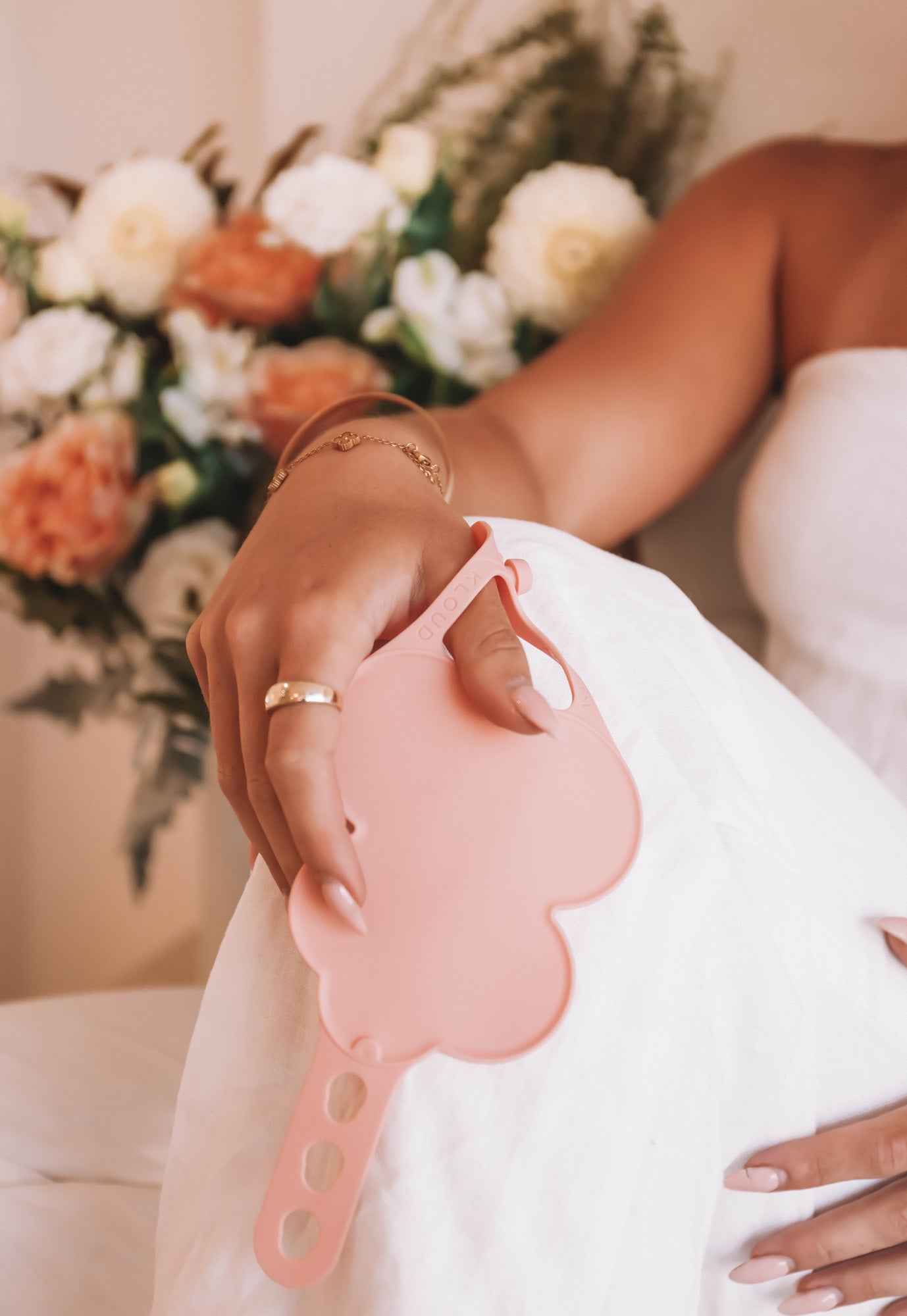 Close-up of a woman's hand resting on her lap, holding a blush pink Kloud Palette with a floral arrangement in the background. This latex-free, wrist-worn tool blends seamlessly into professional makeup setups for weddings, events, or editorial beauty work.