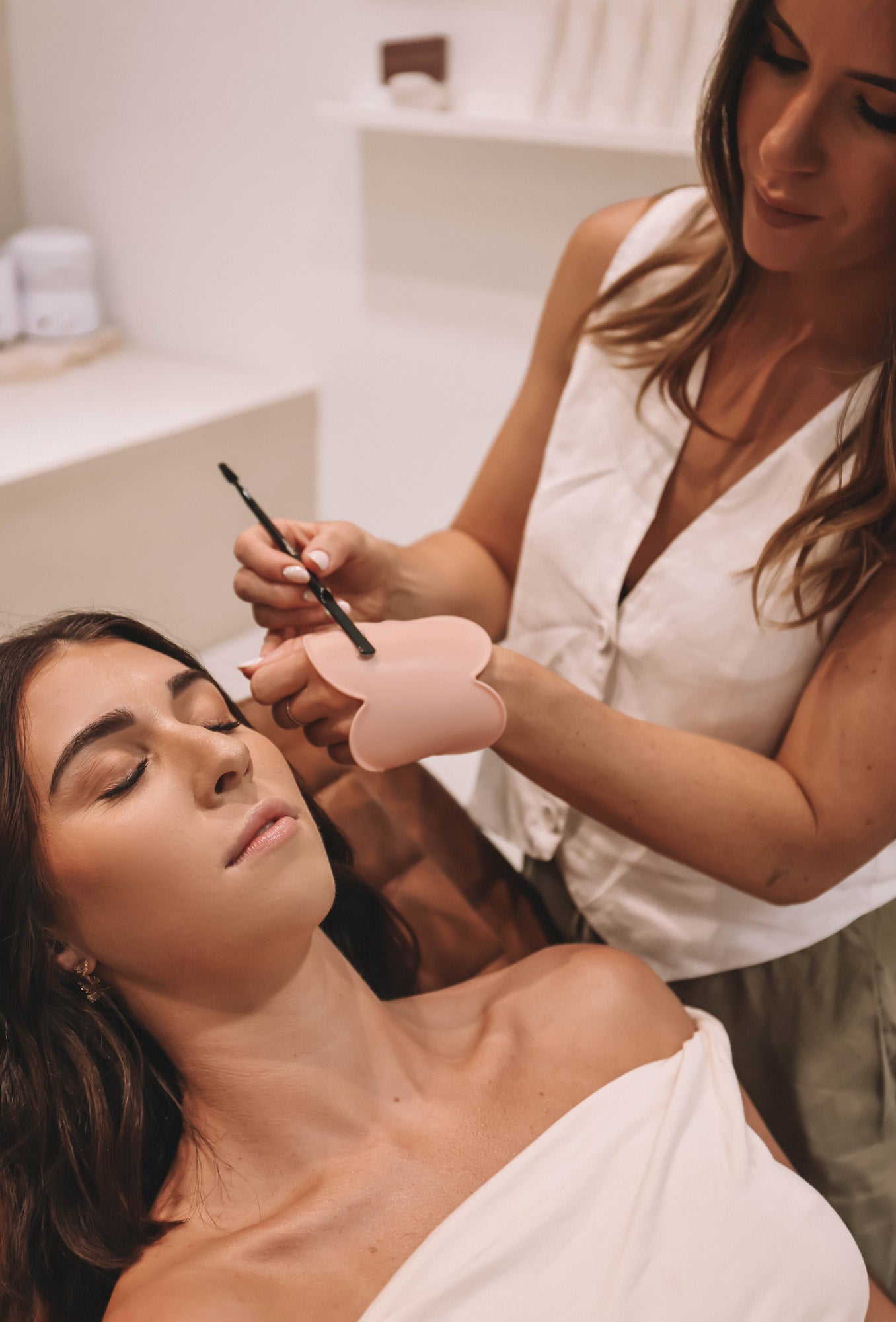 A brow professional performing hybrid dye, lamination, and brow styling on a client while wearing the blush pink Kloud Palette on her wrist. This latex-free palette serves as a professional makeup tool for hygienic, hands-free use during precision beauty treatments.