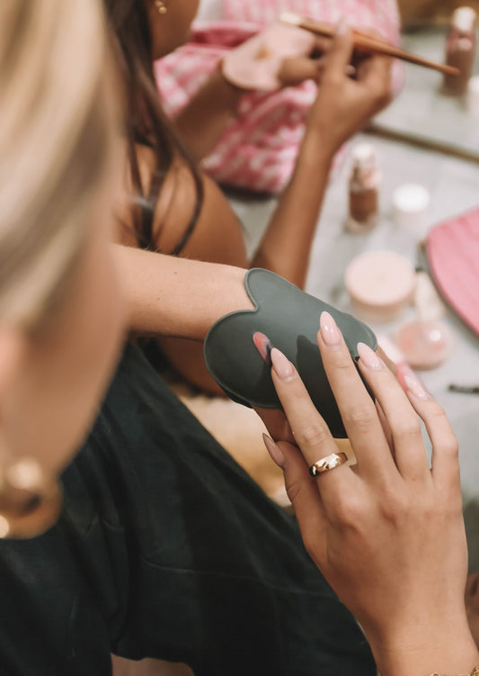 Close-up of a makeup artist using a black Kloud Palette on her wrist to mix cream product, surrounded by beauty tools and soft pink accessories. This latex-free, cloud-shaped palette is a hands-free essential for hygienic and efficient professional makeup application.