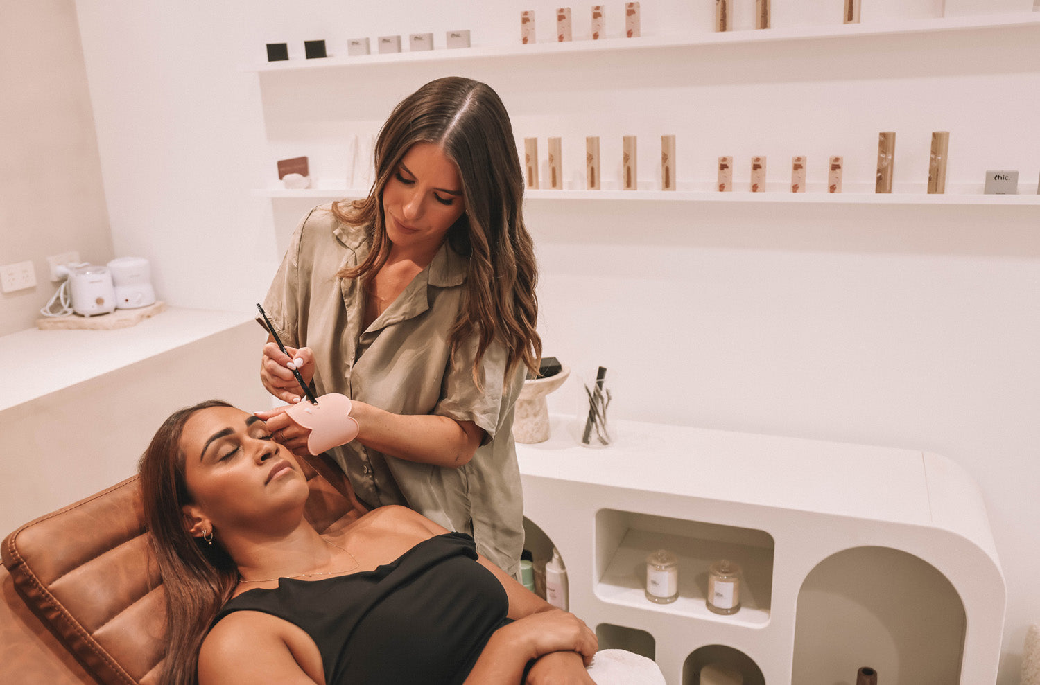 A beauty professional performs a brow service on a client in a modern studio, using the blush pink Kloud Palette worn on her wrist to hold product. This latex-free, cloud-shaped tool supports hygienic, contact-free application—ideal for salon treatments and precision beauty services.