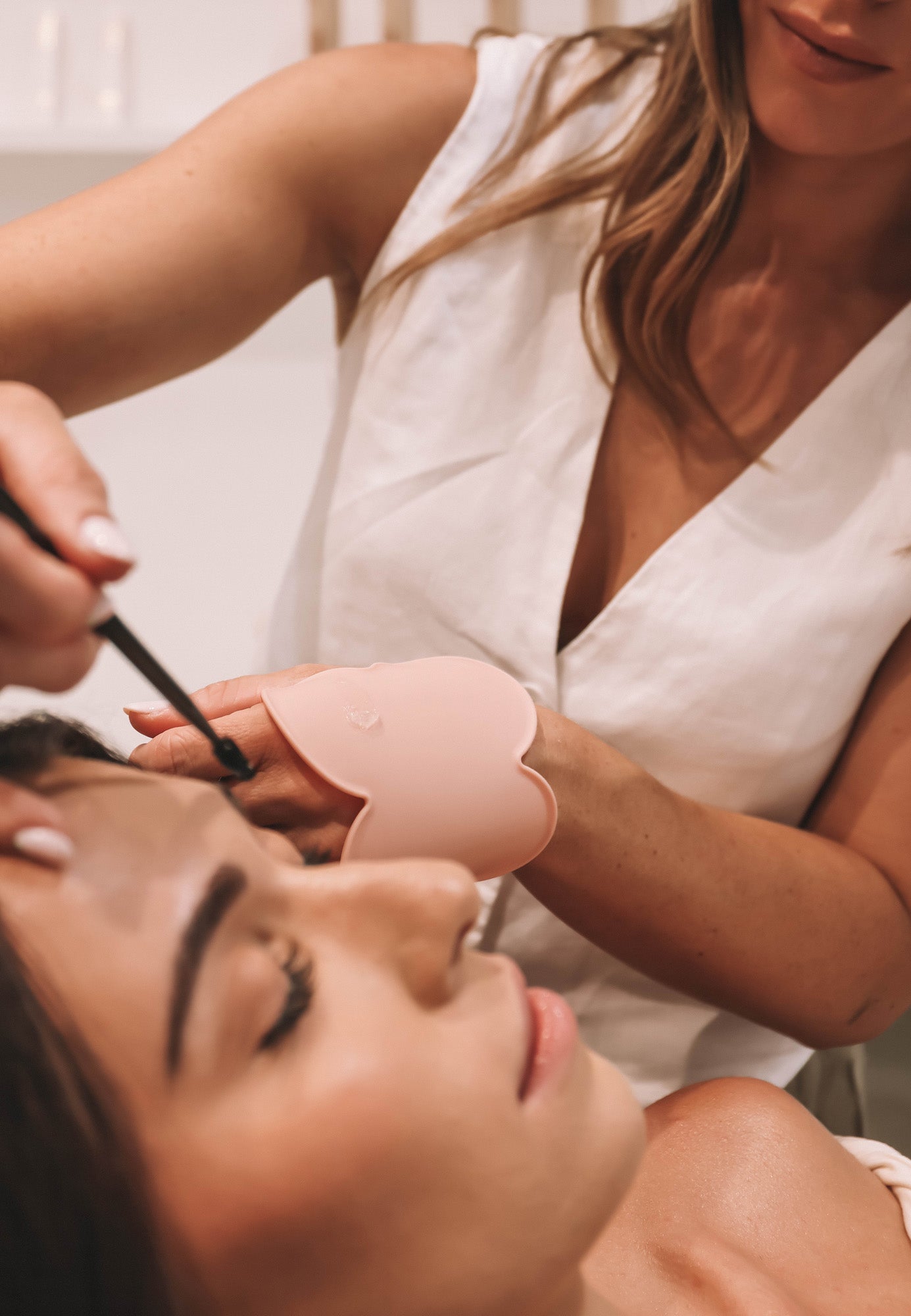 A beauty professional performs precise brow work on a client while wearing the blush pink Kloud Palette on her wrist. This latex-free, cloud-shaped tool provides a clean, skin-safe surface for mixing and accessing product during in-session treatments.