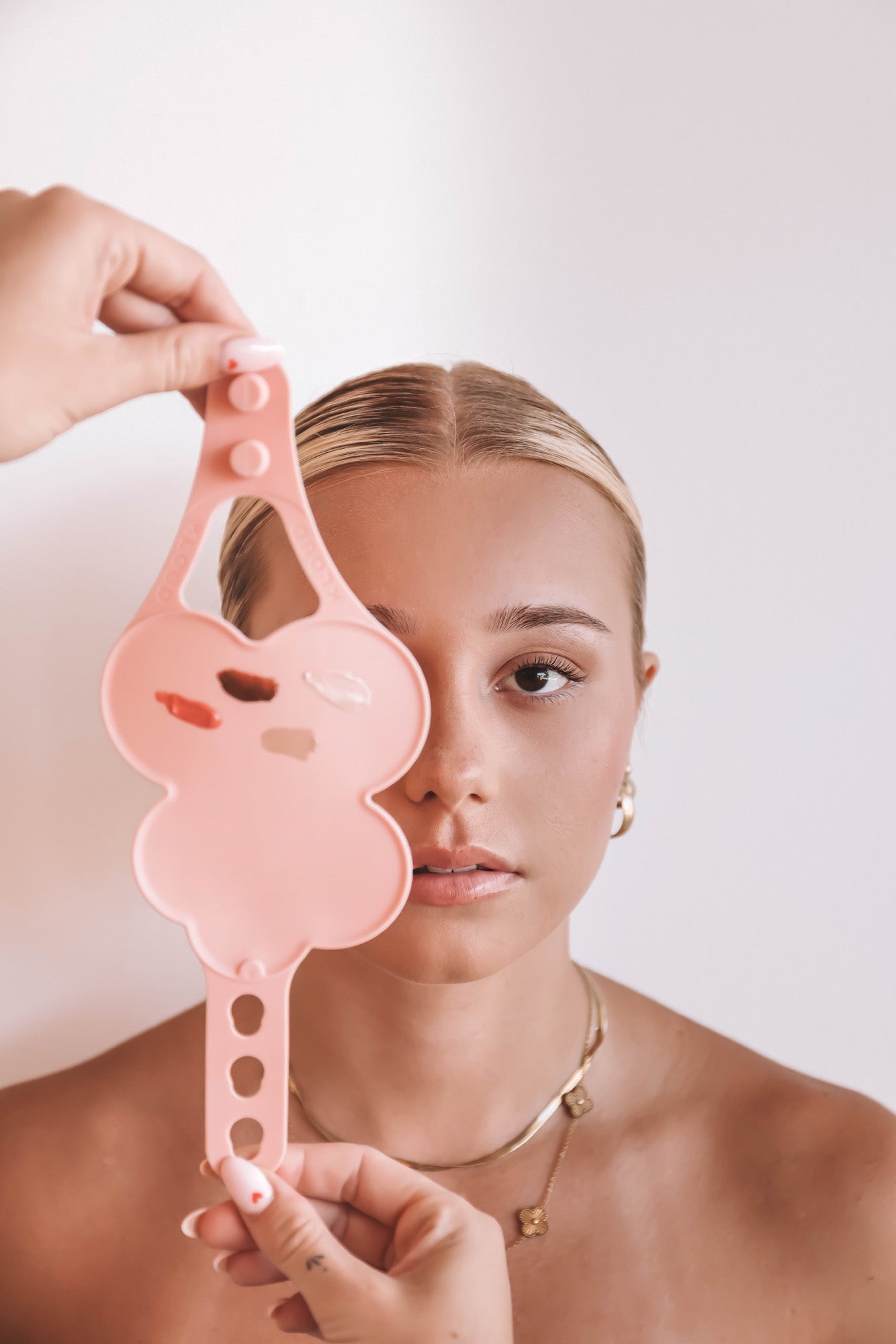 A beauty model holds a blush pink Kloud Palette in front of her face, showcasing four makeup product swatches on its cloud-shaped surface. This professional, latex-free palette is crafted to support contact-free mixing while elevating the artist’s workflow and aesthetic.